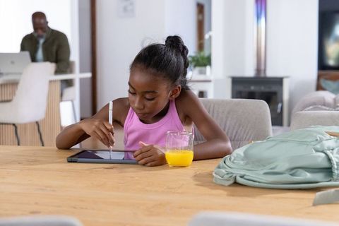 Young black girl using tablet with stylus at kitchen table while parent working remotely