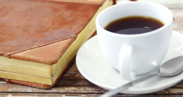 A cup of coffee sits next to an old book on a wooden table, evoking a sense of a relaxing break or a cozy reading time. The scene suggests a moment of tranquility, perfect for those who cherish a quiet coffee break with a good read.