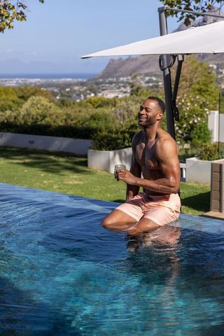 Mature african man relaxing at infinity pool with coastal view holding drink and smiling
