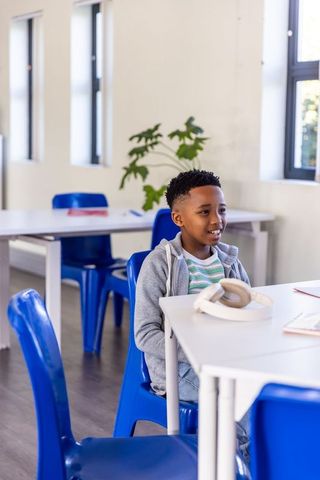 African american boy sitting at classroom table wearing headphones, gray hoodie, learning