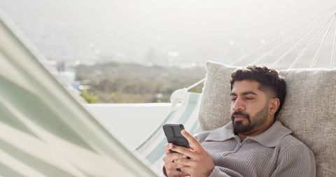 Indian man relaxing on rooftop hammock while scrolling smartphone in knit sweater