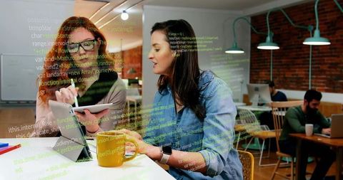 Female developers collaborating over code at coworking counter with tablet and coffee