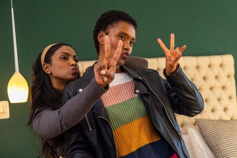 Multicultural female friends posing with peace signs in cozy bedroom wearing leather jacket