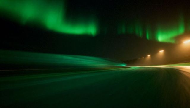 Glowing northern lights reflecting on curving highway with green light trails at night