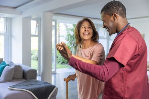 Home care nurse guiding senior woman walking with cane in sunlit living room
