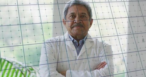 Senior male doctor standing with arms crossed behind glass grid in modern clinic with natural light