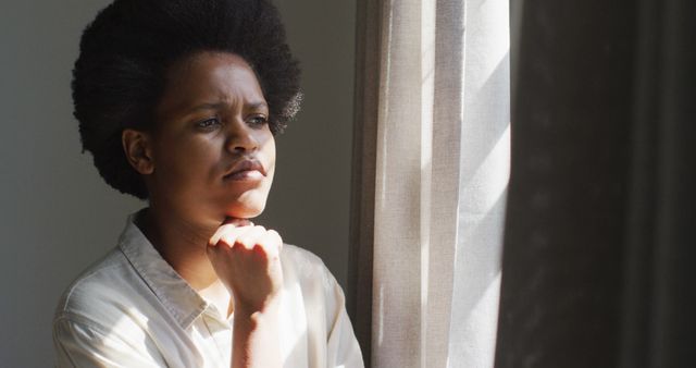 Thoughtful african american woman looking through window in bedroom. domestic lifestyle, spending free time at home.