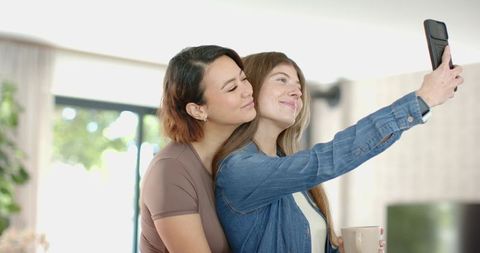 Two women taking selfie while hugging in sunlit living room holding mug and smartphone
