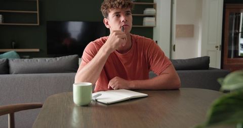 Young man sitting at table thinking with notebook and mug, home study and planning