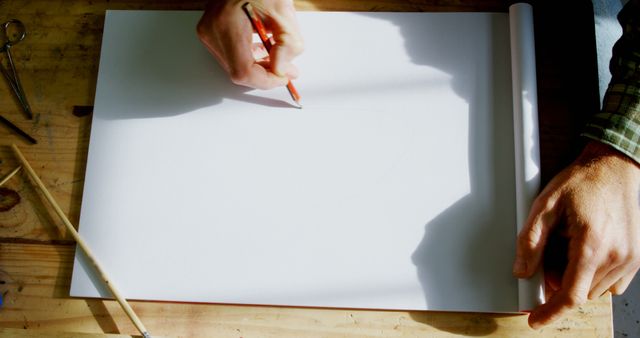A Caucasian person's hand is seen holding a pencil over a blank sheet of paper on a wooden table, with copy space. It suggests the beginning of a creative process, sketching or writing.