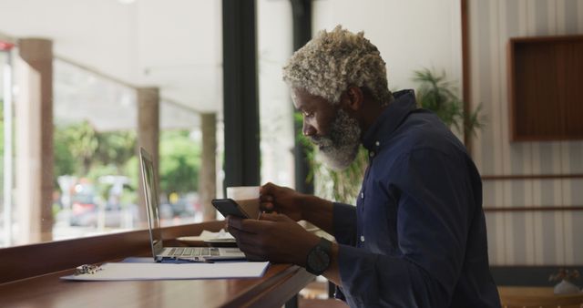 This image shows a mature man working remotely in a cafe. He is using a laptop and a smartphone, demonstrating a modern professional setting. This image can be used for articles or ads about remote work, technology in business, professional lifestyles, or modern work conditions.