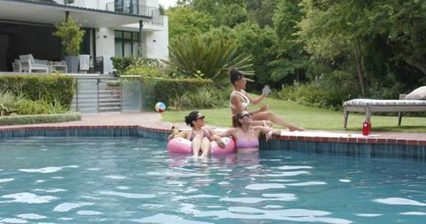 Diverse women lounging on pink inflatable float in backyard pool enjoying sunny summer day