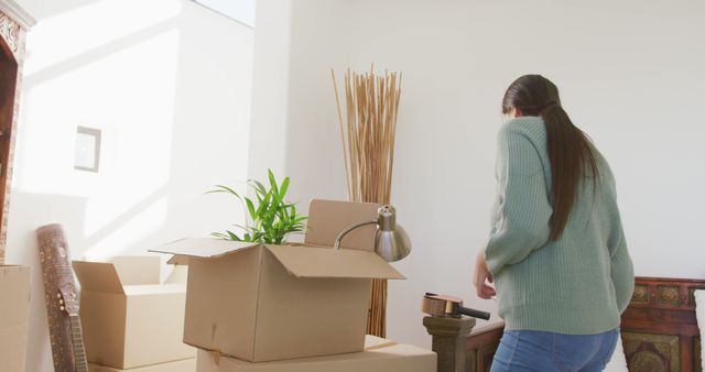 Person packing belongings including house plant into cardboard boxes during the daytime in a well-lit room, suggesting unfolding a new chapter or settling into a new apartment. Useful for illustrating concepts of relocation, changes in living arrangements or lifestyle, and new beginnings. Can be used in real estate advertisements, moving services promotions, or lifestyle blogs discussing home trends and moving tips.