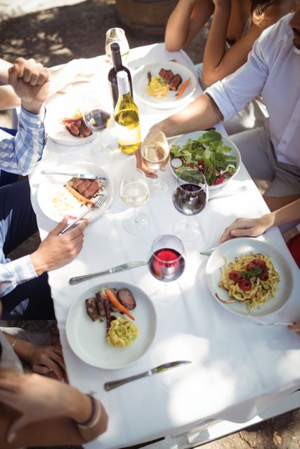 Group of friends enjoying a meal together at an outdoor restaurant. Plates of pasta, salad, and steak are on the table, along with glasses of wine and other drinks. Ideal for use in content related to social gatherings, dining out, food and beverage industry, and lifestyle.