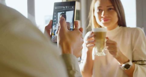Man shooting woman holding layered latte in sunlit cafe with smartphone, candid moment