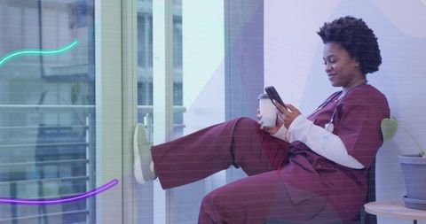 Nurse relaxing on window ledge checking smartphone and holding coffee in clinic lounge