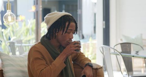 Relaxing african american man sipping coffee on sofa in bright cozy lounge wearing beanie