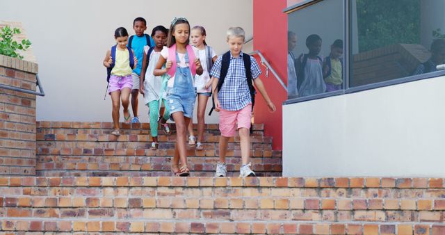 Group of young students walking down brick steps outside a school building, wearing casual clothes and carrying backpacks. Ideal for use in educational campaigns, back-to-school promotions, and articles about school life and childhood development.