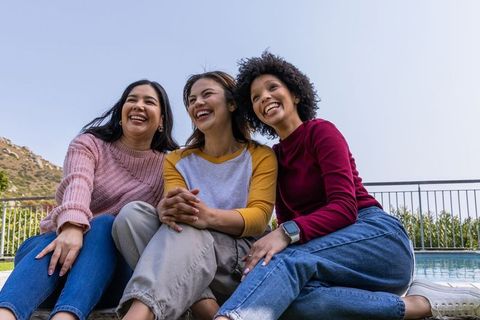 Three diverse friends laughing by pool wearing sweaters and jeans, casual outdoor joy