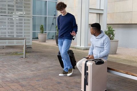 Two young men waiting with suitcases outside modern building chatting and using smartphone