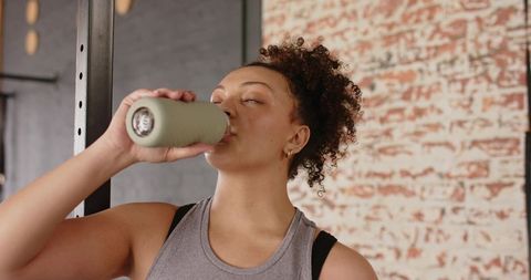 Young woman pausing workout and drinking from reusable bottle in brick-wall gym