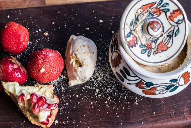 Colorful assortment of strawberries, ice cream, and pomegranate seeds on dark wooden table. Decorative ceramic container adds artistic vibe. Perfect for use in culinary blogs, dessert menus, or rustic kitchen themes.