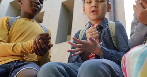 Diverse schoolchildren sitting on steps clapping hands with backpacks and denim jacket