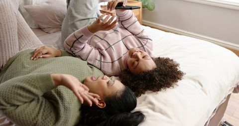 African american friends relaxing on bed sharing smartphone and laughing in daylight