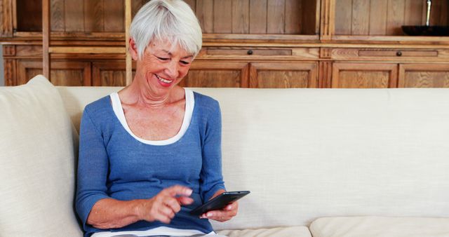 Smiling senior woman sitting on a couch while using a smartphone indoors. Ideal for use in lifestyle, technology, or communication articles, advertisements, and social media promoting modern lifestyles and technology for the elderly.