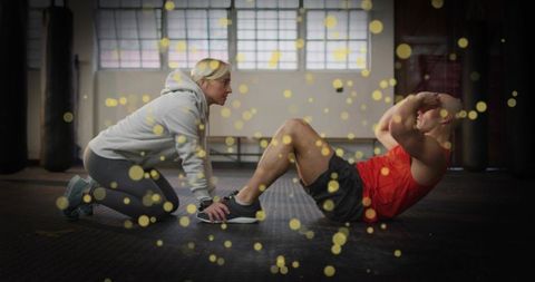 Female trainer holding male athlete feet while performing sit-ups in industrial gym