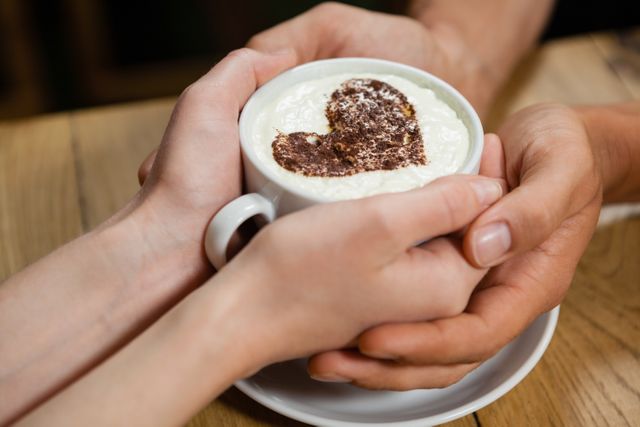 Couple holding a coffee cup with a heart design on the foam, sitting at a wooden table in a cozy cafe. Perfect for use in articles or advertisements about romance, relationships, coffee culture, or cozy cafe settings. Ideal for Valentine's Day promotions, love-themed content, or social media posts about intimate moments.