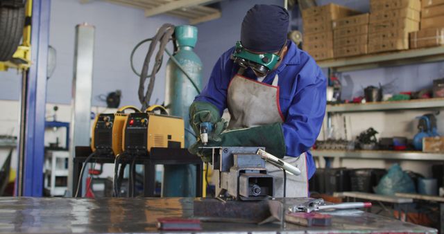 Welder is working on a piece of metal using a welding torch in an industrial workshop. Wearing protective gear including safety goggles and gloves, welder performs precise welding task under safe practices. Shelves stocked with tools and safety equipment are visible in the background. Ideal for depicting industrial work, safety in workplaces, manual labor, and technical skills in engineering environments.