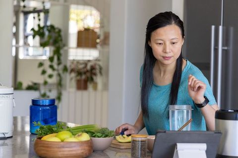 Middle-aged asian woman preparing avocado and greens while following recipe on tablet