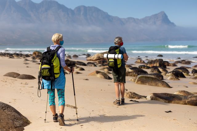 Rear view of caucasian hiker senior couple with backpack and hiking poles walking on the beach. trekking hiking and adventure during coronavirus pandemic concept.