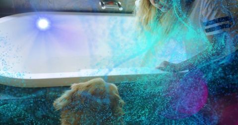 Leaning woman reaching over bathtub to dog with dreamy blue-green light and particles