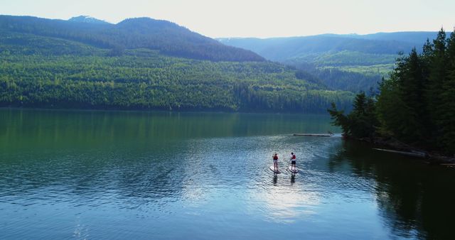 Two people are paddleboarding on a serene lake surrounded by lush green hills, with copy space. Engaging in water sports amidst such tranquil natural beauty offers a refreshing escape from the hustle of daily life.