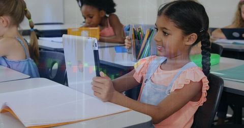 Elementary student holding tablet and learning with notebook and pencils at classroom desk