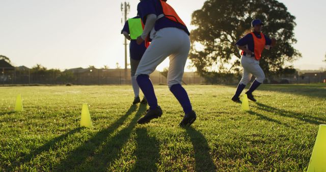 Youth baseball players engage in practice at sunset. They are wearing sports gear and orange training vests, running drills with bright yellow cones set up on a lush green grass field. Ideal for use in articles or promotions about youth sports, teamwork, and fitness, or for illustrating outdoor athletic activities and coaching.