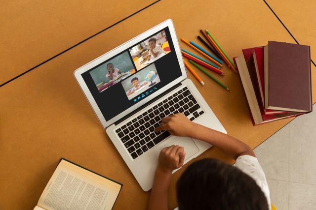 Child participating in remote learning through a virtual classroom on a laptop. Books and colored pencils are on the desk, indicating a learning environment. Ideal for illustrating concepts of modern education, technological integration in learning, and remote classroom setups.