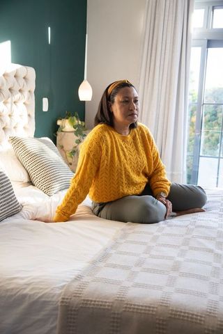 Senior asian woman sitting cross-legged on bed in bright bedroom wearing mustard sweater