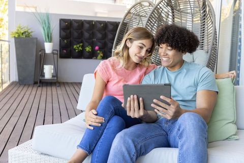Young couple sitting on patio sofa sharing tablet, smiling among modern balcony plants