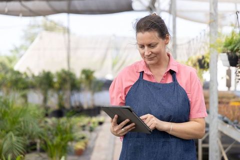 Mature Woman Checking Plant Inventory on Tablet in Greenhouse Aisle