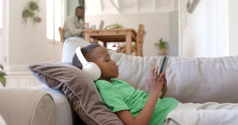 Boy relaxing on sofa wearing headphones using tablet, casual at-home screen time