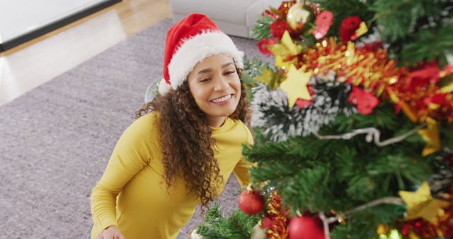 Image of happy biracial woman in santa hat smiling and decorating christmas tree at home. Christmas, celebration, tradition, happiness and inclusivity concept.