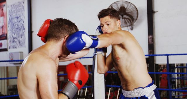 Two boxers engage in sparring in a gym boxing ring. One boxer wearing blue gloves lands a punch on the other's head, who is wearing red gloves. The action looks competitive and intense, highlighting the physical fitness and focus required in the sport of boxing. This image is ideal for use in articles about fitness, boxing training techniques, athletic training, or sports competition. It can also be used to promote boxing gyms or fitness centers.
