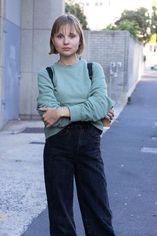 Young woman standing on sidewalk holding coffee cup and backpack wearing green sweater
