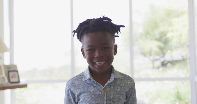 Image shows a smiling African American boy with dreadlocks standing in a bright room. He is wearing casual clothing and appears cheerful. The background is softly lit with natural light coming through large windows. Perfect for use in advertisements related to children's products, family life, joyful moments, and home interiors.