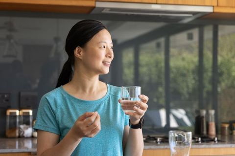 Asian woman taking pill holding glass of water in modern kitchen morning wellness routine