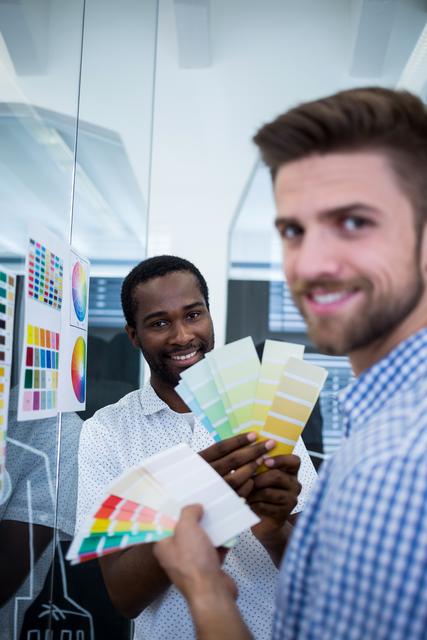 Two male graphic designers are holding color swatches while working together in a modern office. They are smiling and appear to be collaborating on a project, making decisions about color schemes. This image can be used to depict teamwork, creativity, and the design process in a professional setting. Ideal for articles, blogs, or advertisements related to graphic design, creative industries, and office environments.