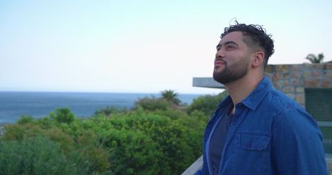 Contemplative man gazing toward sea horizon from coastal balcony with greenery